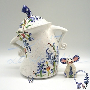 Puede incluir: Un juego de tetera y figura de ratón de cerámica blanca. La tetera presenta diseños florales azules y una tapa a juego. La figura del ratón tiene grandes orejas rosas y azules y sostiene una flor azul. El juego está pintado a mano.