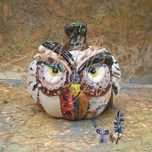 May include: A ceramic owl-shaped bottle stopper. The owl has large yellow eyes, a patchwork design of red, brown, and blue, and a green top. The background is a textured, multi-colored surface.