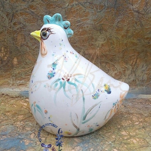 May include: White ceramic chicken figurine with a blue comb and wattle. The chicken is decorated with a floral pattern in shades of green, blue, and yellow.