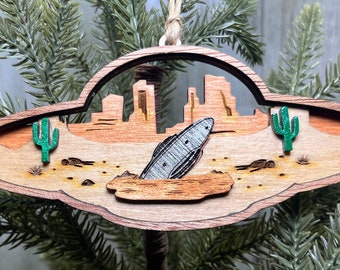 May include: A wooden Christmas bauble shaped like a flying saucer with a crashed spaceship inside. The bauble is decorated with a desert landscape, including cacti and a rocky terrain.