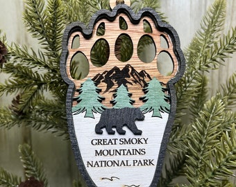 May include: Wooden ornament shaped like a bear paw print with a silhouette of a bear and mountains inside. The ornament is decorated with green trees and the text "Great Smoky Mountains National Park".