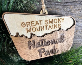 May include: A wooden ornament shaped like the outline of the Great Smoky Mountains National Park. The ornament is brown with a light wood grain and has the words "Great Smoky Mountains National Park" engraved on it.