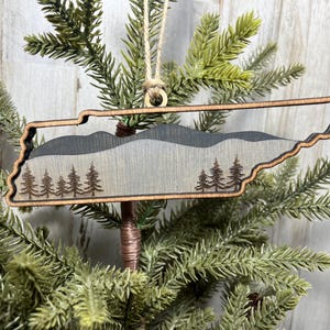 May include: Wooden ornament shaped like the state of Tennessee with a mountain range and pine trees design. The ornament is painted in shades of gray and brown.