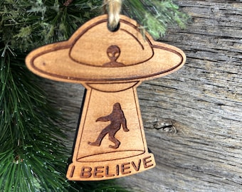 May include: Wooden Christmas ornament shaped like a UFO with an alien inside and a Bigfoot walking underneath. The ornament has the words "I BELIEVE" written on the bottom.