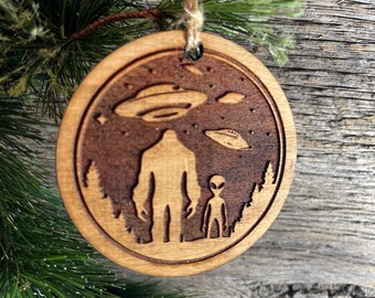 May include: A round wooden Christmas ornament with a laser-etched design of Bigfoot and an alien standing in a forest with three flying saucers in the sky.