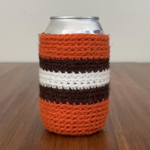 May include: A knitted can cooler in orange, brown, and white stripes. The cozy is wrapped around a silver aluminum can. The handmade item is designed to keep beverages cold and is a unique accessory for any occasion.