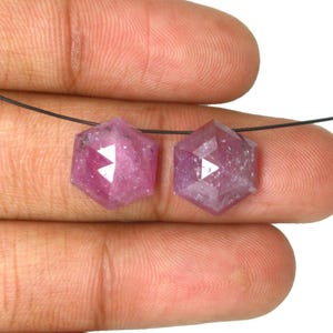 May include: Two hexagonal, rose-cut pink gemstones with a faceted surface, strung on a thin black thread. The stones exhibit a translucent, deep magenta hue with natural internal inclusions and a polished finish, showcasing a geometric shape.