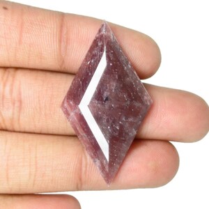 Puede incluir: Una gema facetada en forma de diamante, de color rojo púrpura. La gema presenta un diseño geométrico blanco en el centro. La piedra se sostiene en una mano sobre un fondo blanco.