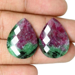 Może przedstawiać: Dwa kamienie szlachetne w kształcie łezki o fasetowanej powierzchni. Kamienie szlachetne charakteryzują się żywą mieszanką kolorów, w tym głębokiej czerwieni, zieleni i bieli. Kamienie są trzymane w dłoni, prezentując ich wypolerowany wygląd.