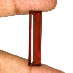 May include: A long, rectangular, deep red gemstone with a faceted surface. The stone features a clear, polished finish and a vibrant, saturated crimson hue, showcasing sharp, straight edges and a slender, elongated baguette-like shape.