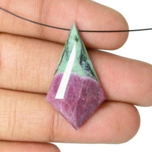 May include: A gemstone pendant with a diamond shape. The pendant features a vibrant mix of colors, including deep magenta, green, and black. A white line runs down the center. The pendant is suspended from a thin black cord.