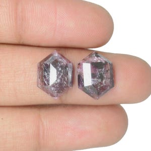 Könnte beinhalten: Zwei sechseckig geschliffene, facettierte Edelsteine mit einem gräulich-violetten Farbton. Die Edelsteine haben ein kristallines Aussehen mit inneren Einschlüssen, präsentiert vor neutralem Hintergrund. Geeignet für die Schmuckherstellung.