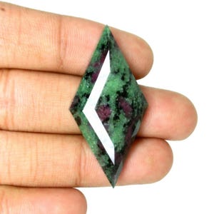 Op de afbeelding: Een ruitvormige edelsteen met een groene basiskleur, zwarte en rode insluitsels. Het oppervlak is gefacetteerd en gepolijst, waardoor het licht reflecteert. De steen wordt in een hand tegen een witte achtergrond gehouden.