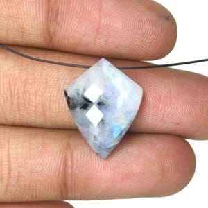 May include: A kite-shaped moonstone gemstone featuring a faceted surface with a translucent white appearance, subtle blue iridescence, and dark inclusions. The stone is drilled at the top and suspended on a thin black string for display.