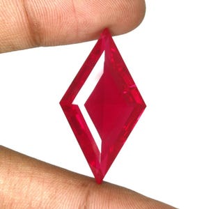 May include: A vibrant, translucent red gemstone cut into a sharp, elongated diamond or kite shape. The stone features precise, faceted edges that create a geometric pattern with a central rhombus-shaped facet, reflecting light across its polished surface.