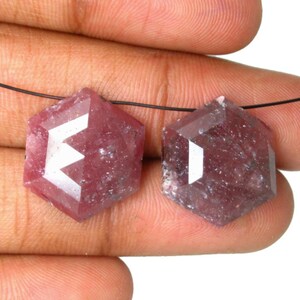 Peut inclure: Deux pierres précieuses hexagonales de couleur rose avec une surface à facettes. Chaque pierre présente un dessus plat et poli ainsi qu'une forme géométrique distincte, enfilées sur un fil noir fin, révélant une teinte rose-rouge translucide.