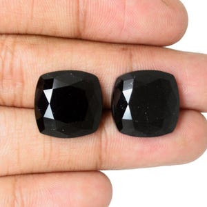 May include: Two loose, cushion-cut black gemstones with a faceted surface. The stones have a square shape with rounded corners and a high-gloss, reflective finish. Each gemstone displays a deep, opaque black colour with sharp, geometric facets.