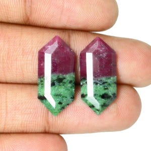 Puede incluir: Dos gemas facetadas, cada una con una parte superior roja oscura y una base verde. Las gemas se sostienen en una mano, mostrando sus superficies pulidas y su combinación de colores única. Las gemas tienen forma hexagonal.