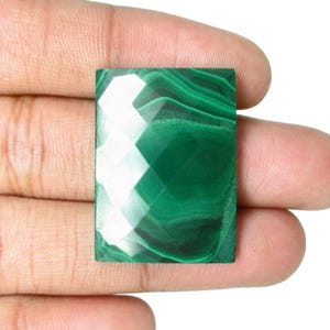Può includere: Una gemma di malachite rettangolare e sfaccettata, di un verde vibrante e con una superficie lucida. La gemma è tenuta in una mano, mostrando i suoi intricati motivi e la sua bellezza naturale. Le sfaccettature della pietra riflettono la luce, creando un effetto scintillante.