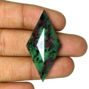Puede incluir: Una piedra preciosa facetada en forma de diamante con un patrón moteado de inclusiones de color verde oscuro y rojo oscuro. La piedra tiene una superficie pulida y reflectante con bordes afilados y simétricos, mostrando una mezcla de texturas minerales opacas y semitranslúcidas.