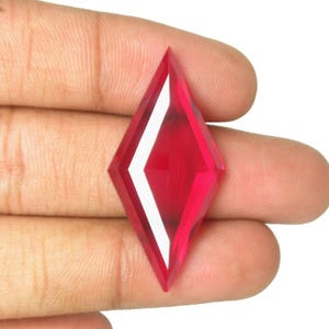 May include: A vibrant, translucent red gemstone cut into a sharp, elongated diamond or marquise shape. The stone features multiple faceted surfaces that create distinct geometric reflections and highlights across its deep crimson, polished body.