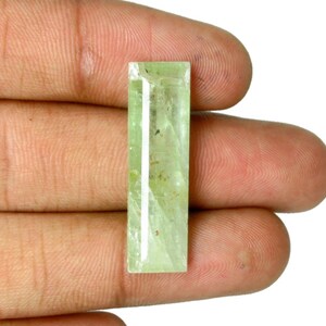 May include: A rectangular, faceted gemstone with a pale, translucent light green hue. The stone features a smooth, polished surface with visible internal inclusions and a clear, elongated shape that reflects light across its flat top facets.