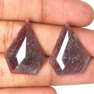May include: Two loose, shield-shaped gemstones with a deep reddish-purple hue and a faceted surface. The stones exhibit a translucent quality with internal inclusions and a polished finish, reflecting light across their geometric, multi-sided cut.
