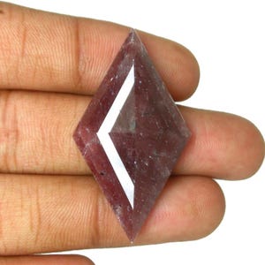 May include: A kite-shaped, faceted gemstone with a deep reddish-purple hue and a translucent, slightly cloudy appearance. The stone features a sharp, geometric diamond-like silhouette with a prominent, bright white light reflection across its surface.