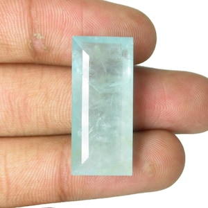Può includere: Una pietra preziosa rettangolare sfaccettata di colore blu acquamarina pallido e traslucido. La pietra presenta una superficie superiore piatta con bordi smussati e una struttura cristallina chiara che lascia passare la luce attraverso le faccette.