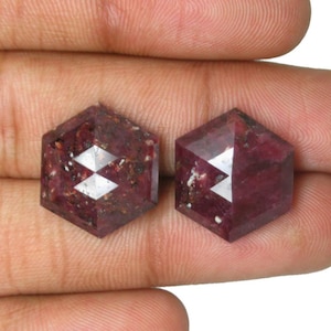 Puede incluir: Dos piedras preciosas de color rojo intenso con corte hexagonal, inclusiones oscuras visibles y superficie facetada. Las piedras muestran un tono burdeos oscuro con motas minerales naturales en toda la estructura, con una forma geométrica y acabado pulido.