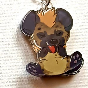 Puede incluir: Un llavero de hiena con un cierre dorado. La hiena tiene un cuerpo marrón con manchas oscuras, orejas negras y una lengua roja. Un accesorio divertido.