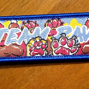 Pode incluir: Um porta-chaves retangular bordado com uma borda azul e um anel de metal. O design apresenta um fundo amarelo e azul com várias pegadas de patas castanhas e cor-de-rosa. O texto "TEAM PAWS" está bordado em azul claro no centro.