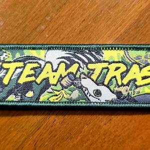 Pode incluir: Um porta-chaves retangular bordado com uma borda verde escura e um anel de metal. O design apresenta um fundo amarelo com o texto TEAM TRASH em letras amarelas em negrito, acompanhado por ilustrações de um guaxinim e uma criatura.