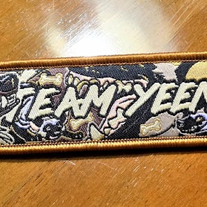 Pode incluir: Um porta-chaves retangular bordado com uma borda dourada e um anel de metal prateado. O design apresenta ilustrações de hienas e o texto "TEAM YEEN" numa fonte clara sobre um fundo escuro estampado com motivos de ossos.