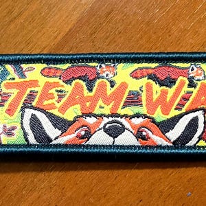 May include: A rectangular fabric keyring with a dark green border and a silver metal ring. The design features multiple red pandas against a yellow and green background with the text TEAM WAH printed in bold orange letters across the centre.