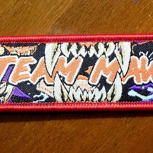 May include: A rectangular embroidered keychain with a red border and a black background featuring a graphic of sharp teeth and the orange text "TEAM MAWS" in the center. A silver metal split ring is attached to the left side of the fabric tag.