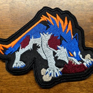 May include: An embroidered patch featuring a stylised wolf creature with blue fur, orange spikes along its back, and white underbelly fur. The creature is depicted in a crouching position, biting into a dark red object with visible bone-coloured details.