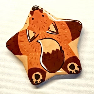 Può includere: Un oggetto decorativo a forma di stella con un disegno di volpe dei cartoni animati. La volpe è raffigurata in tonalità di marrone, arancione e beige, con una coda con la punta bianca. L'oggetto ha una finitura lucida, probabilmente in ceramica o simile.