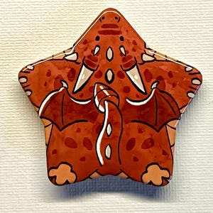 Peut inclure: Figurine de dragon en céramique en forme d'étoile, dans les tons orange, marron et blanc. Le dragon a des ailes, des cornes et une longue queue. La figurine est peinte à la main avec une finition brillante. Le design est fantaisiste et de style cartoon.
