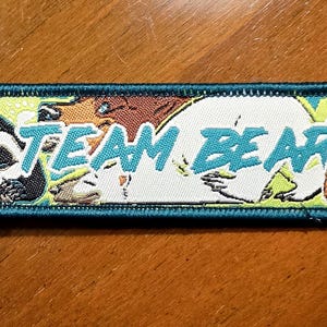 Puede incluir: Un llavero rectangular bordado con un borde verde azulado oscuro que muestra un panda, un oso polar y un oso pardo. El texto "TEAM BEAR" está impreso en azul claro en el centro del parche, el cual está unido a una anilla de metal plateado.