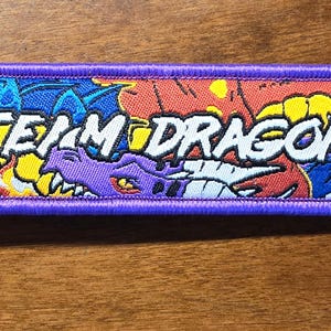 Può includere: Un portachiavi rettangolare ricamato con un bordo viola e un anello in metallo. Il design presenta illustrazioni di draghi colorati in blu, rosso e viola con accenti gialli. Il testo "TEAM DRAGON" è ricamato in bianco al centro.