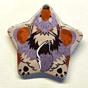 May include: Star-shaped pin badge with a cartoon fox design. The badge has a light purple background with orange and grey accents. The fox has a black and white tail and paws. The badge is likely ceramic or enamel.