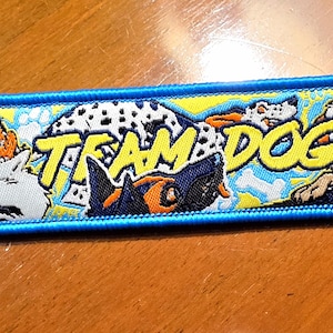 Pode incluir: Um porta-chaves retangular bordado com uma borda azul brilhante e um fundo amarelo com várias ilustrações de cães e o texto TEAM DOG em letras amarelas em negrito. Um anel de metal prateado está preso no lado esquerdo.