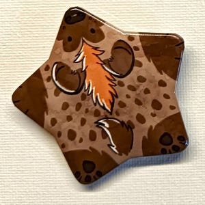 Può includere: Un elemento decorativo a forma di stella, marrone e beige, con un disegno di animali dei cartoni animati. Il disegno presenta un motivo maculato marrone, con un accento arancione e bianco. Il pezzo ha una finitura lucida.