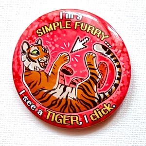 Può includere: Un bottone rotondo rosso con un'illustrazione di una tigre dei cartoni animati. La tigre è arancione e nera, con occhi verdi, ed è sdraiata sulla schiena. Il testo sul bottone recita "I'm a SIMPLE FURRY... I see a TIGER I click."
