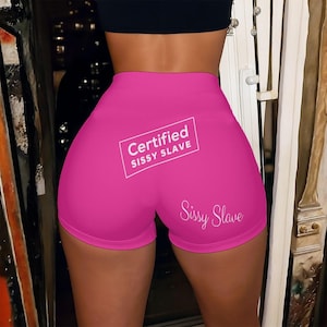 May include: Hot pink athletic shorts with the text "Certified Sissy Slave" in a white rectangular box on the back. Below the box is the cursive text "Sissy Slave".