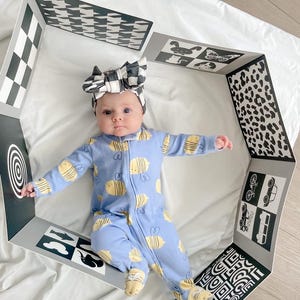 Puede incluir: Un beb&eacute; con una diadema negra y blanca y un mono azul y amarillo con estampado de abejas est&aacute; tumbado dentro de una alfombra de juego de patrones negros y blancos. La alfombra tiene diferentes patrones e im&aacute;genes, incluyendo un patr&oacute;n a cuadros, un patr&oacute;n de pata de gallo, un estampado de leopardo y un objetivo.