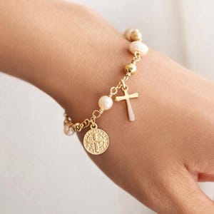 May include: Gold-coloured bracelet with a cross charm, a round pendant, and white pearls. The bracelet is on a wrist, highlighting the delicate design and religious theme. The bracelet is a piece of jewellery.