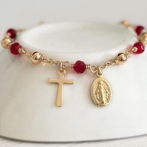May include: Gold-toned rosary bracelet featuring red beads and gold accents. The bracelet includes a cross charm and a medallion with a religious figure. The bracelet is displayed on a white surface.
