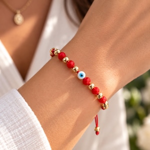 Op de afbeelding: Een kralenarmband met afwisselend ronde rode kralen en metallic goudkleurige bollen, in het midden voorzien van een enkele wit-blauw-zwarte boze oog kraal. De armband is geregen aan een dun rood koord met verstelbare uiteinden.